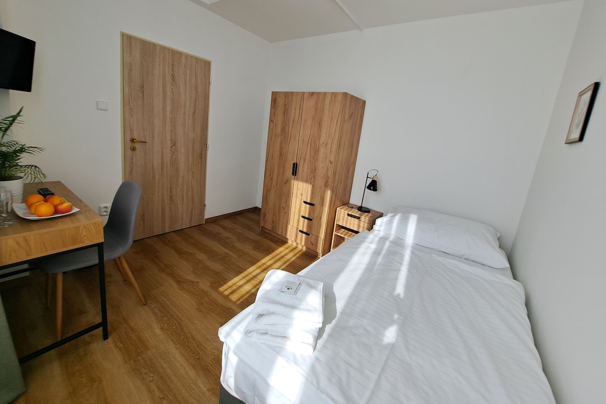 Et lille, godt oplyst rum med et træskrivebord, en stol, en seng med hvide sengetøj og en stor garderobe.