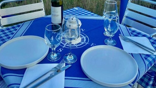 Mesa dispuesta con platos, vasos y una botella, con vista a una playa y barcos.