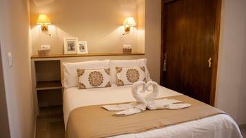Habitación de hotel con una cama, dos lámparas fijadas a la pared, una puerta de madera y una pequeña estantería con cuadros enmarcados.