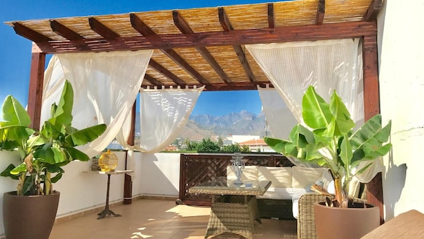 Un espacio de estar al aire libre con techo y vistas a montañas, con una pérgola de madera, cortinas blancas y plantas en macetas.
