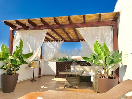 A covered outdoor seating area with a view of mountains, featuring a wooden pergola, white curtains, and potted plants.