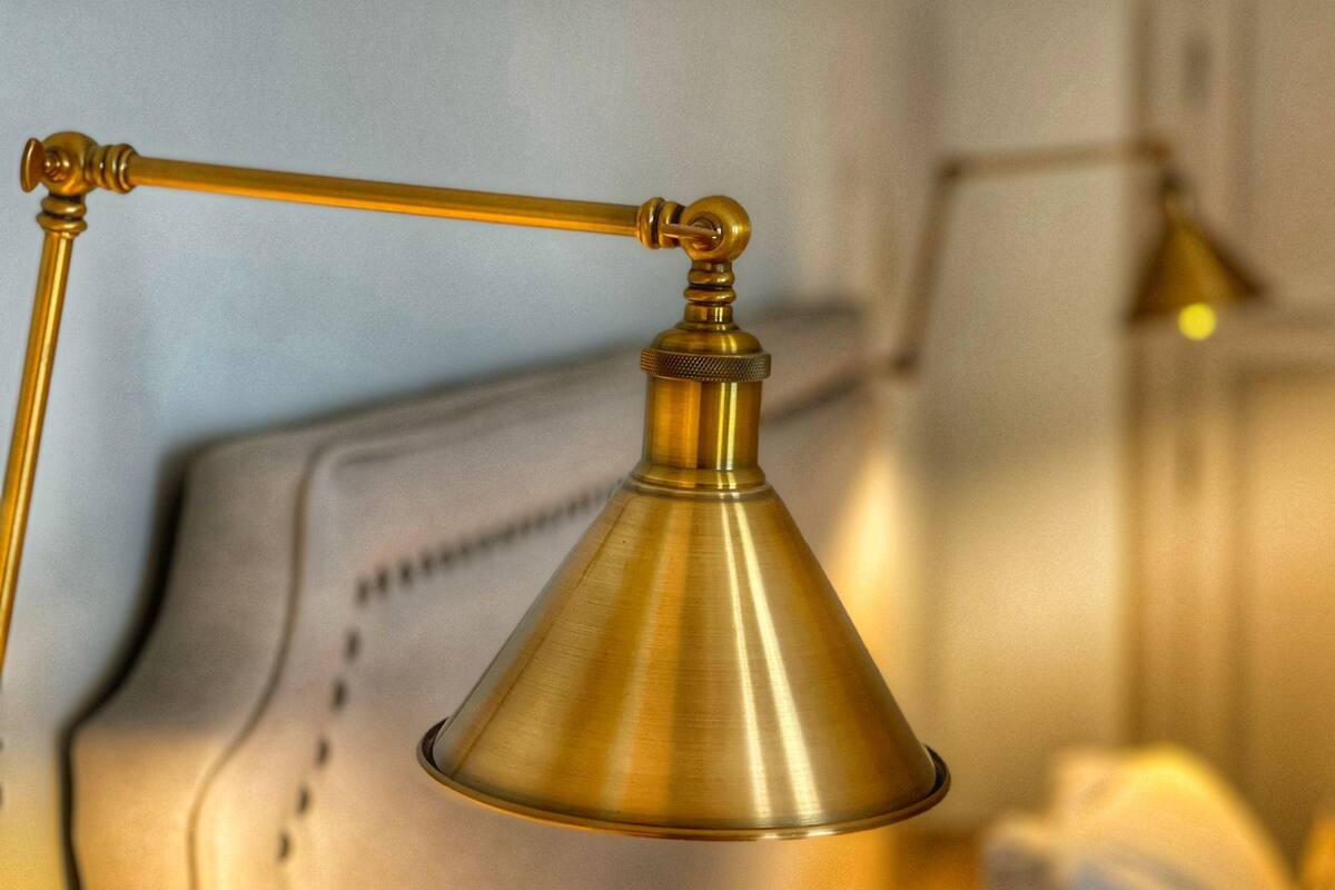 A brass desk lamp with a conical shade, mounted on a wall with a white, padded headboard.