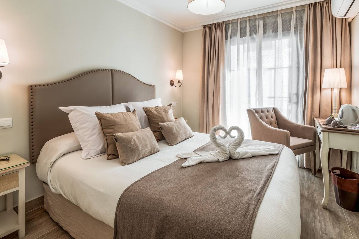 A neatly made bed with a swan-shaped towel, a chair, a desk with a lamp, and a window with curtains.
