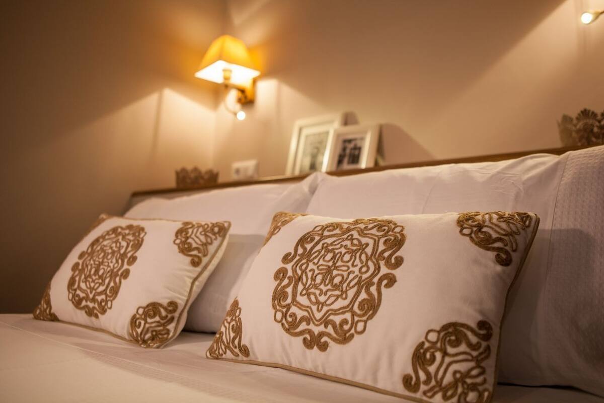 A bed with two pillows featuring intricate gold embroidery, a wall-mounted light fixture, and a framed picture on the wall.