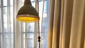 Un sillón con brazos y patas de madera, con tapizado texturizado, ubicado frente a una ventana con cortinas translúcidas y una lámpara de pie con pantalla estampada.