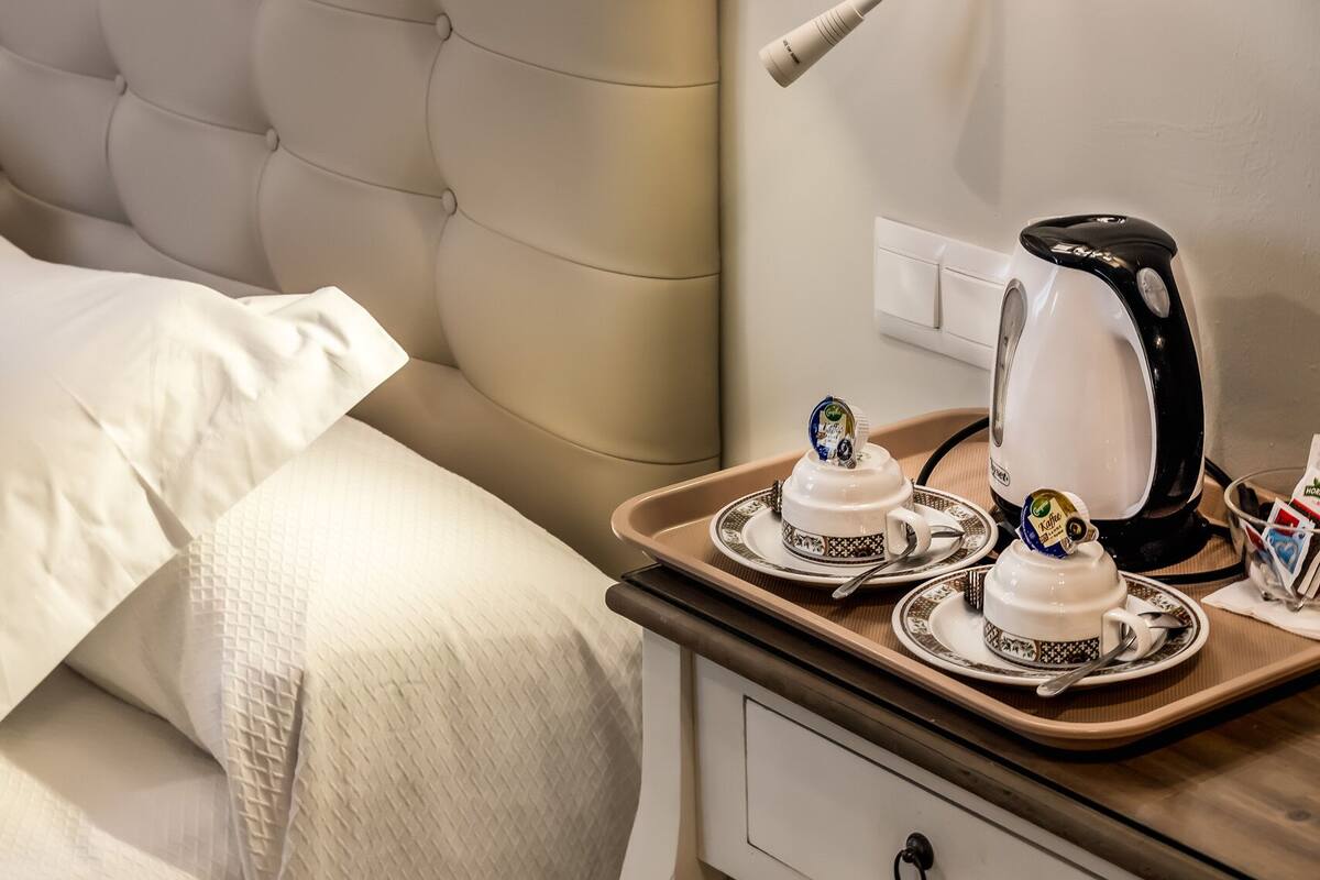 A neatly made bed with a tufted headboard, a bedside table with a kettle, teapot, and cups, and a wall-mounted light fixture.