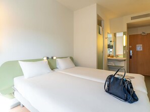 A hotel room with a bed, a nightstand, a mirror, a door, and a handbag.