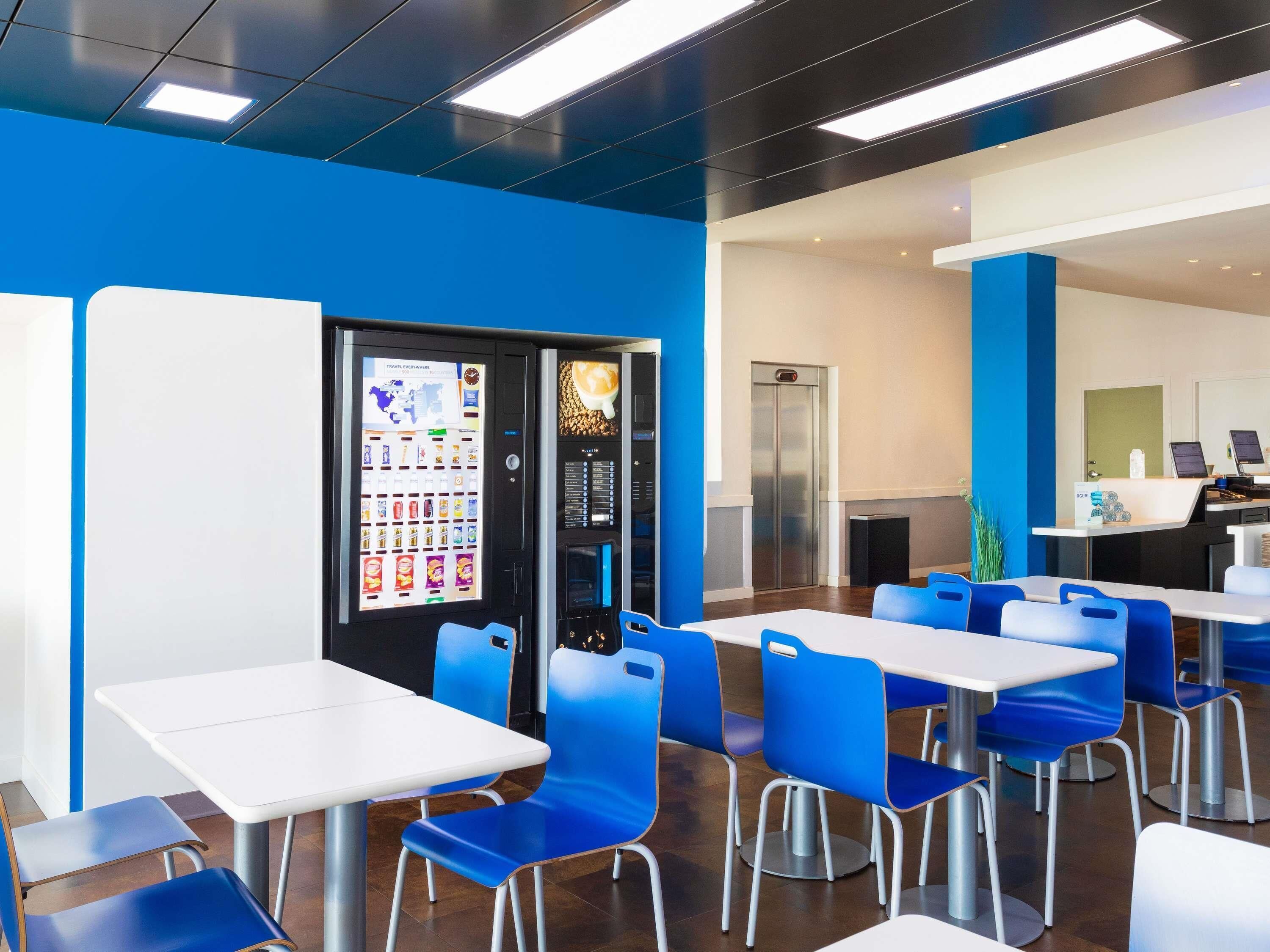 A modern dining area with blue chairs and white tables, a vending machine, and a small elevator.