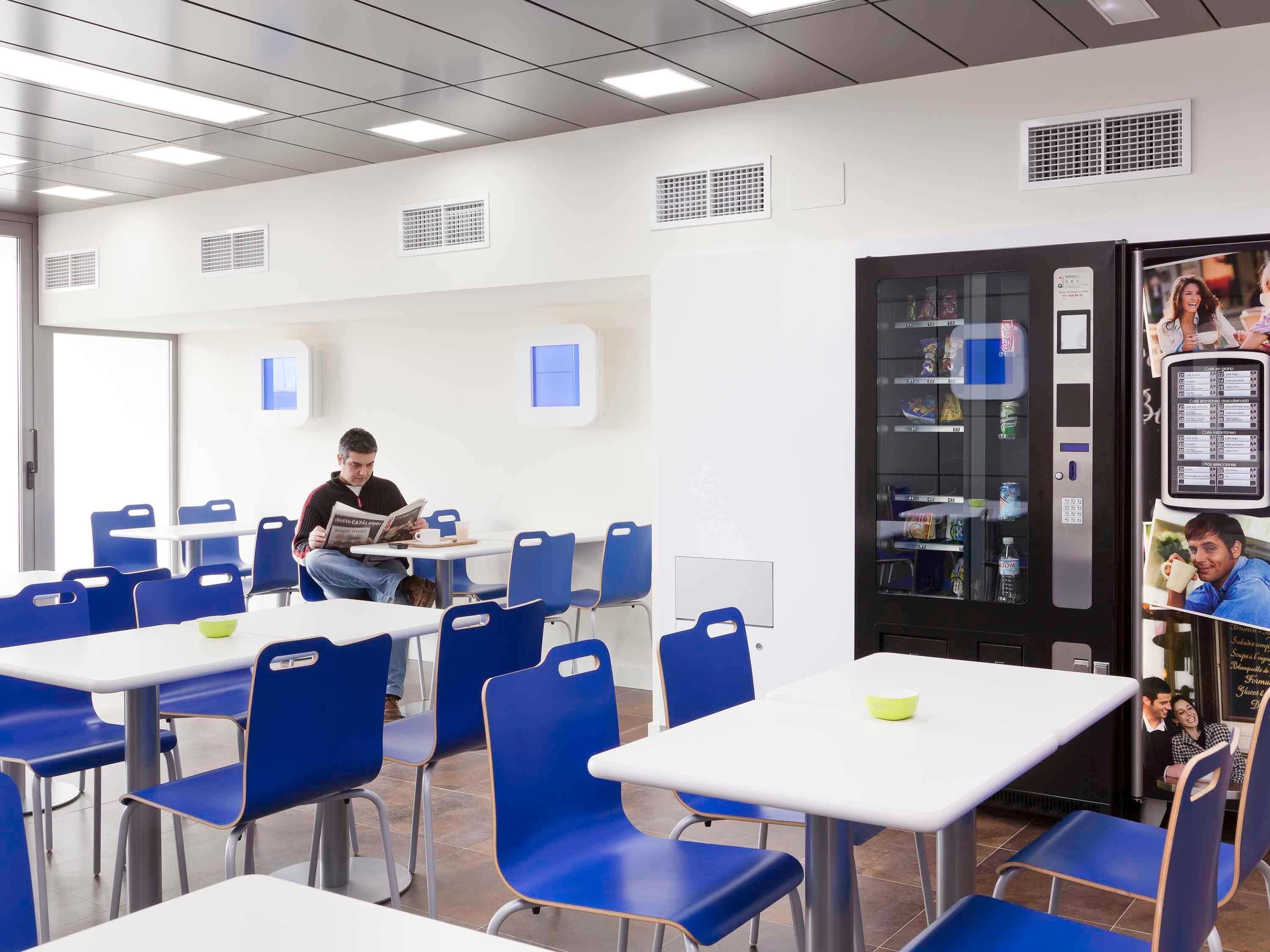 Un café moderno con sillas azules, mesas blancas y una máquina expendedora.