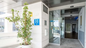 Entrada de un edificio moderno con una planta en maceta, un letrero azul con la letra 'H' y una puerta de cristal que da al interior.