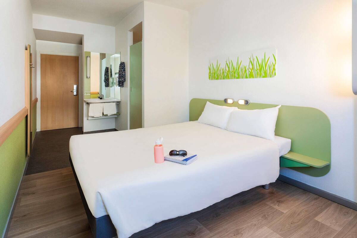 Habitación de hotel con cama, mesita de noche, espejo, pared verde y suelo de madera.
