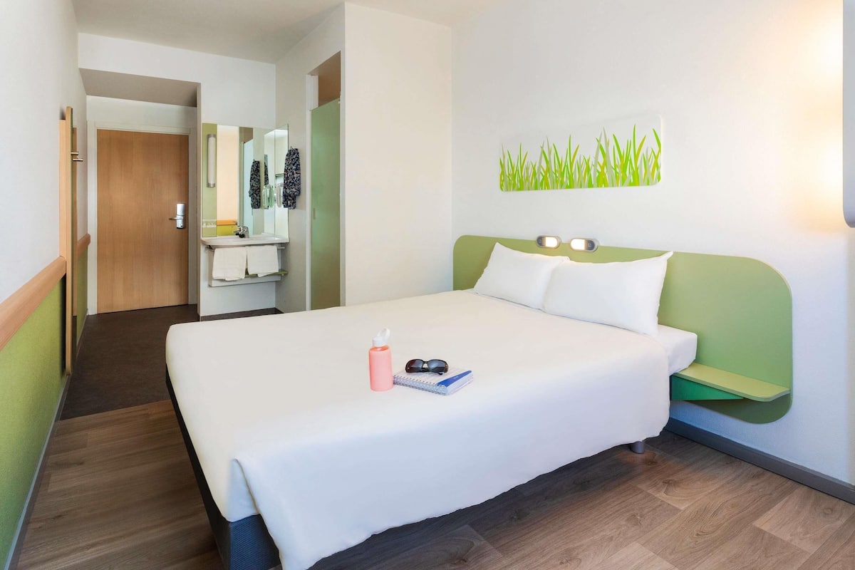 Habitación de hotel con cama, mesita de noche, espejo, pared verde y suelo de madera.