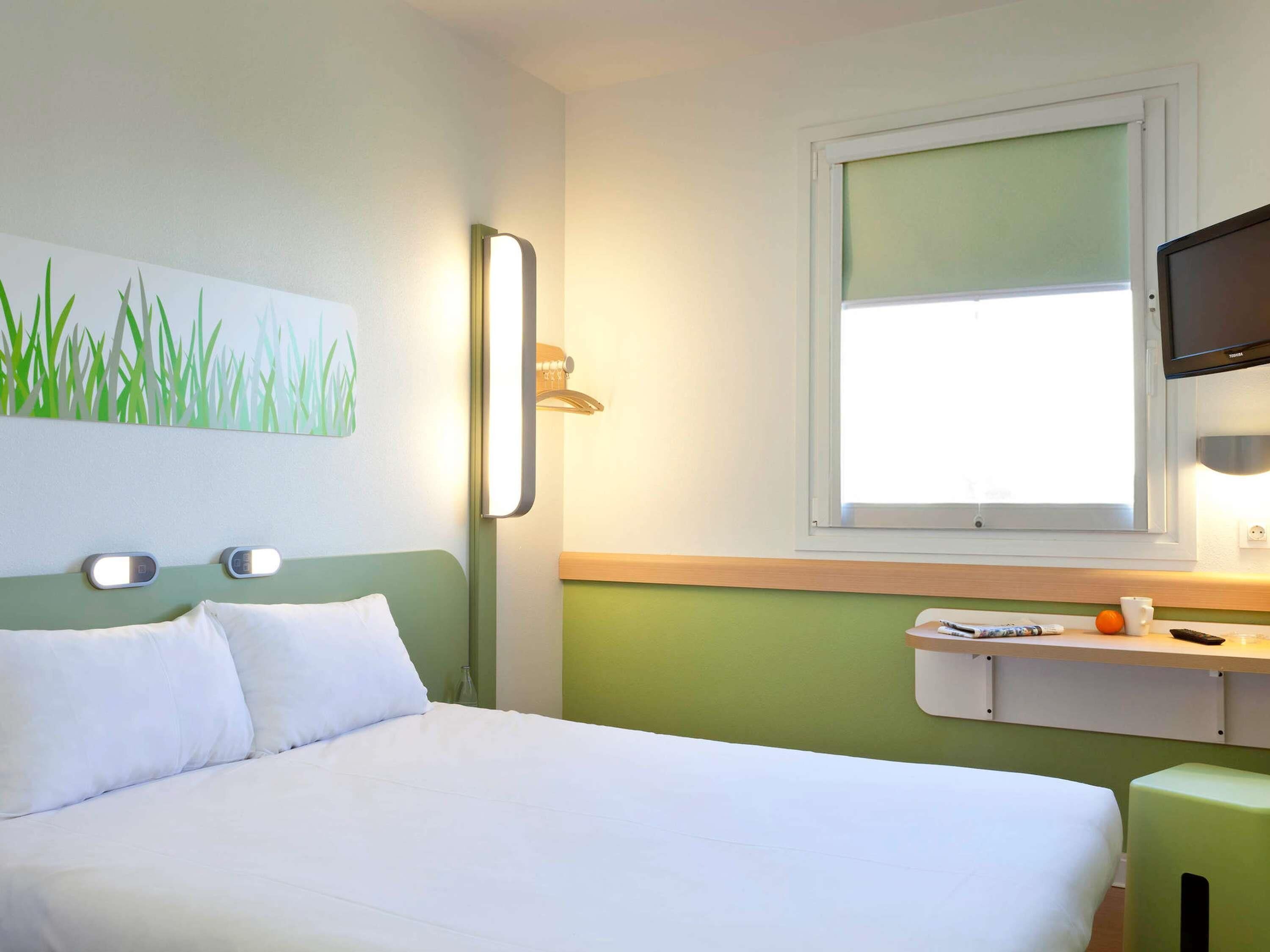 A hotel room with a bed, a painting of grass on the wall, a bedside table with a cup and a remote control, and a window with a view.