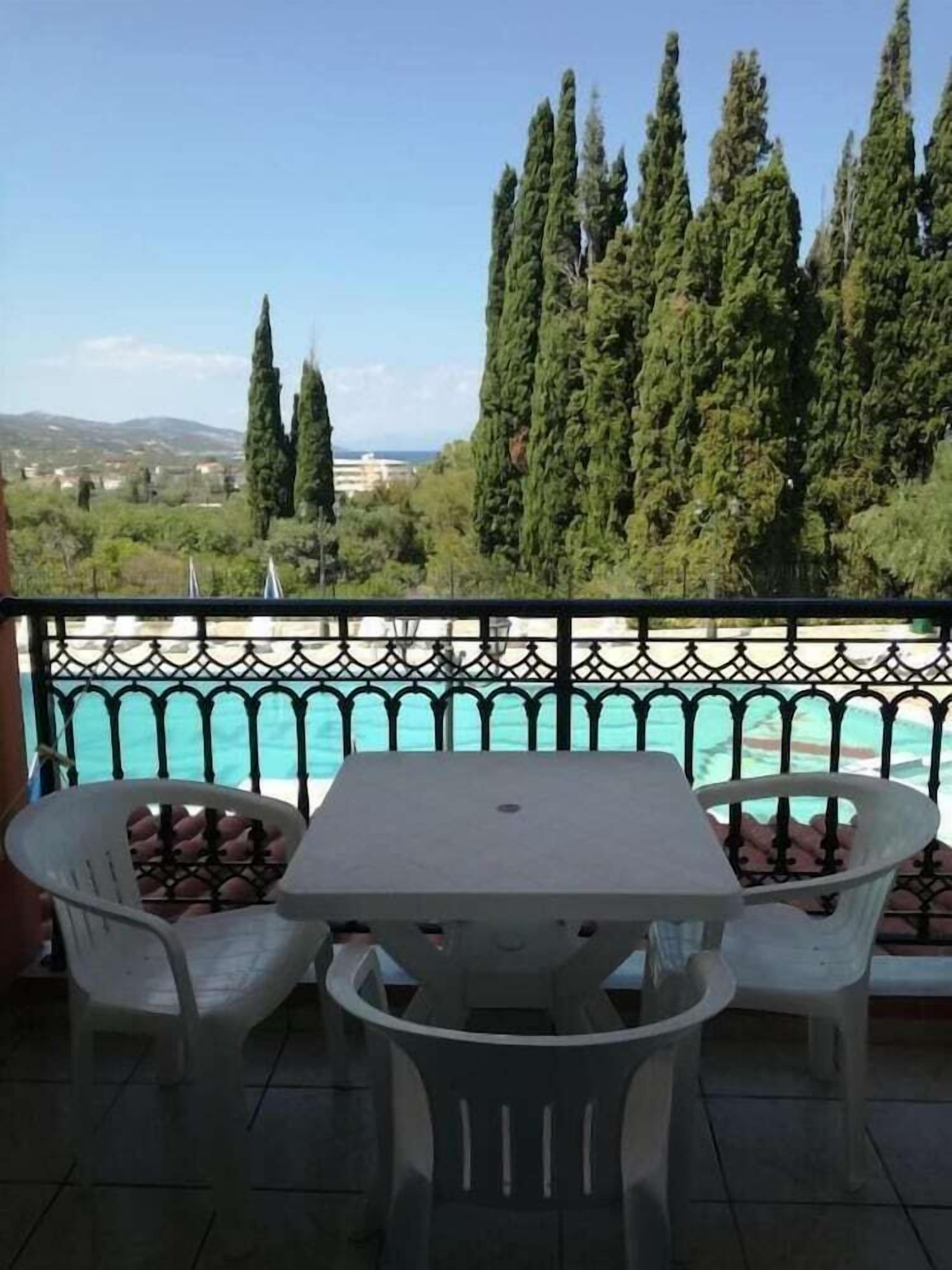A balcony with a table and chairs, overlooking a pool and a view of trees and buildings.