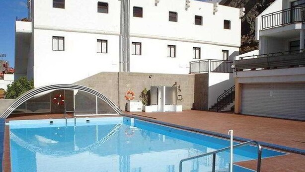 Una piscina con cerramiento de vidrio, rodeada por una solana con baldosas y un edificio blanco con múltiples ventanas y balcones.