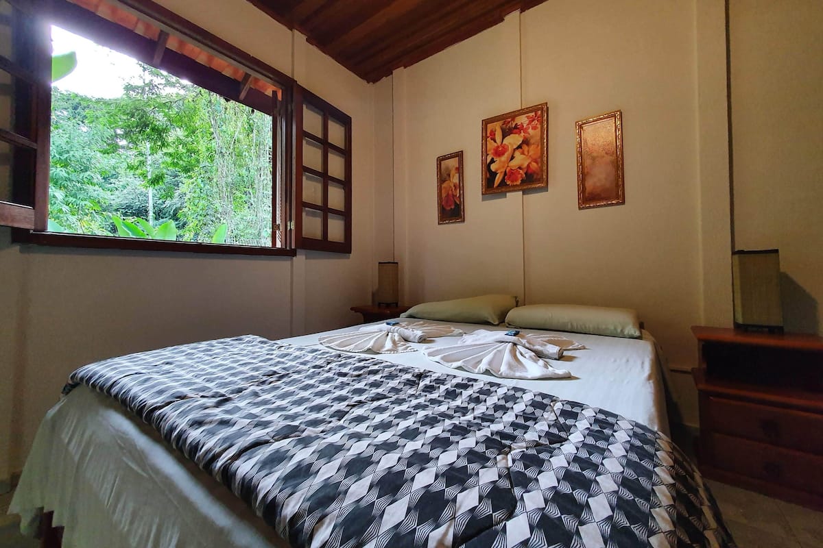 Quarto com uma cama, janela com vista para o verde e três quadros na parede.