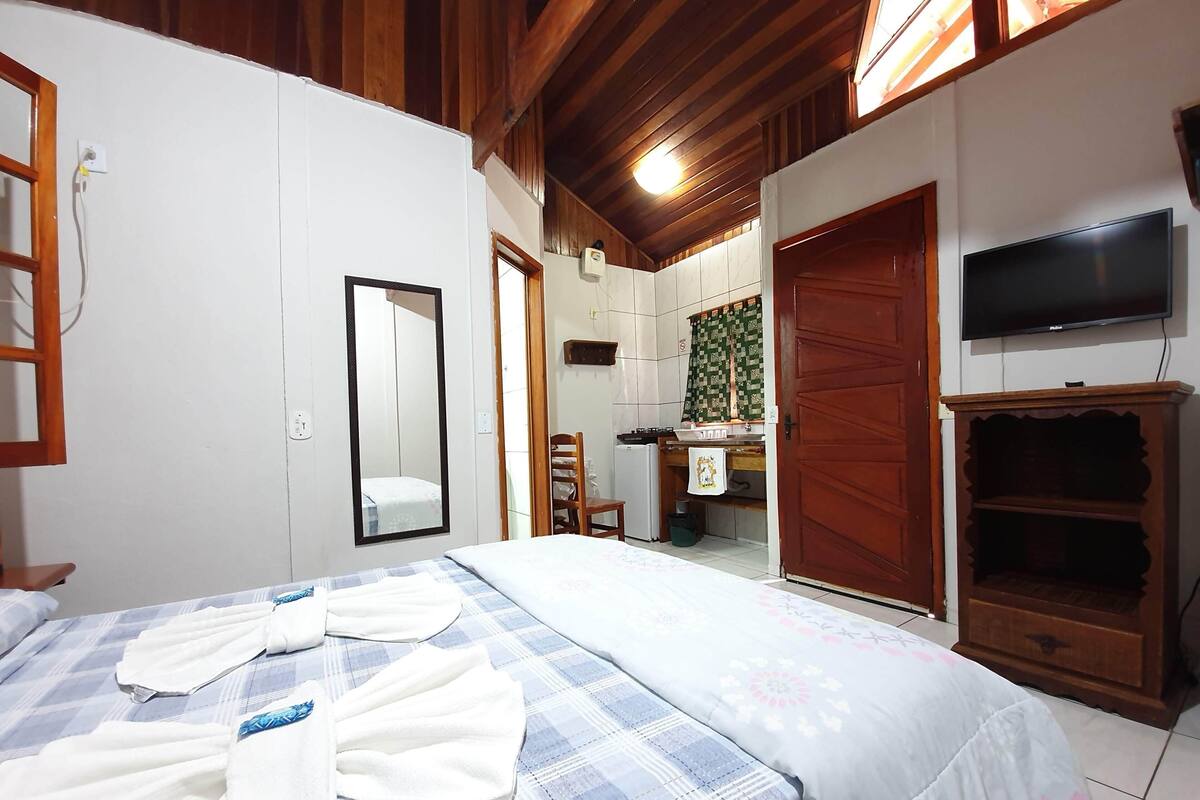 Quarto de hotel com cama, televisão, guarda-roupa de madeira, espelho e banheiro visível por uma porta aberta.
