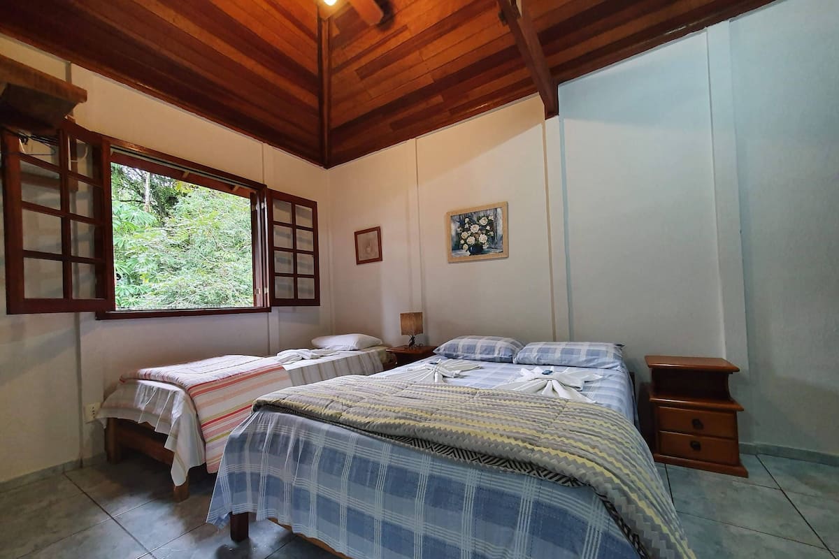 Quarto com duas camas, janela com persianas de madeira, um quadro na parede e uma pequena mesa de cabeceira de madeira.