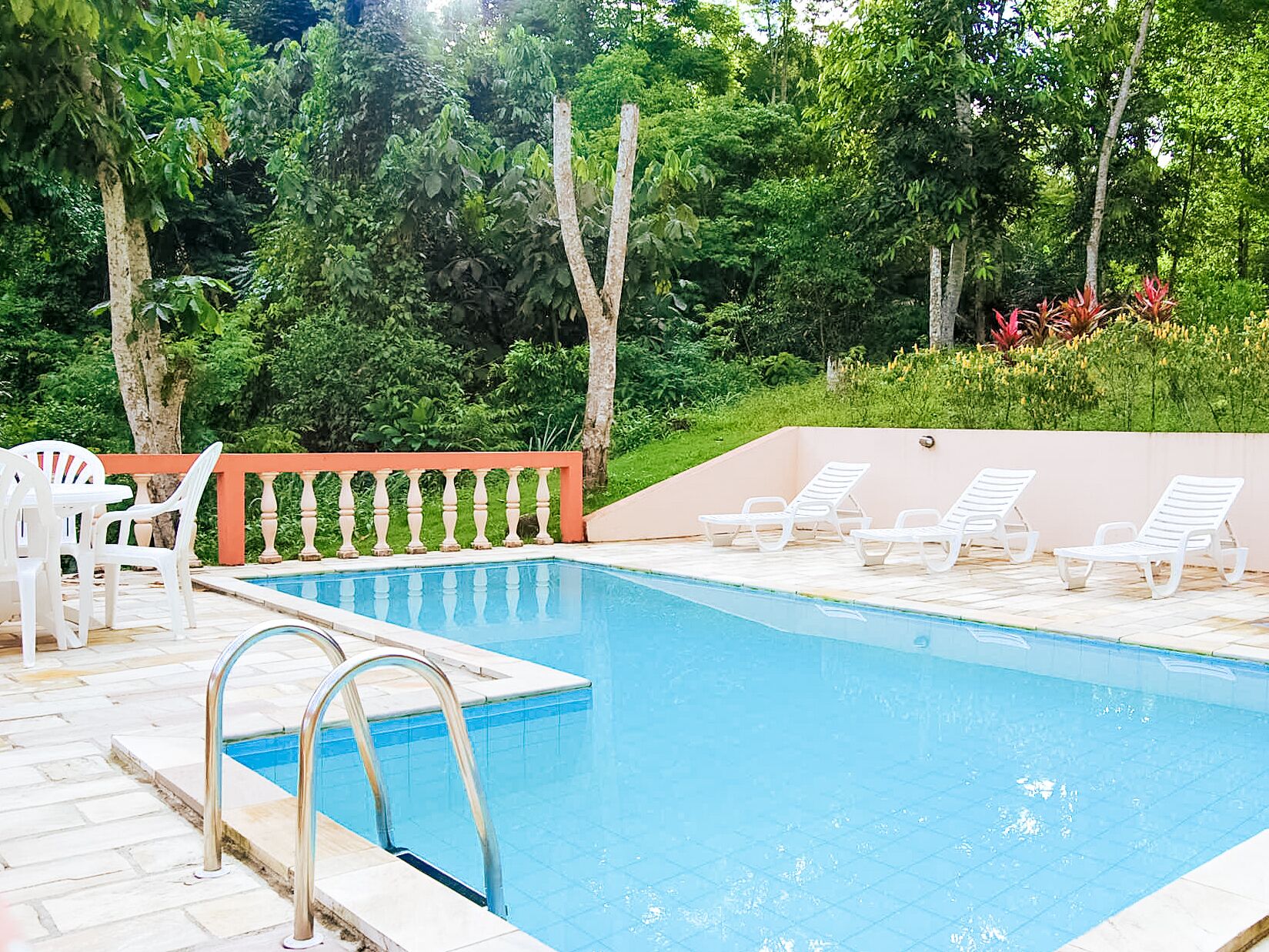 A swimming pool with a tiled deck, surrounded by lush greenery and white lounge chairs.
