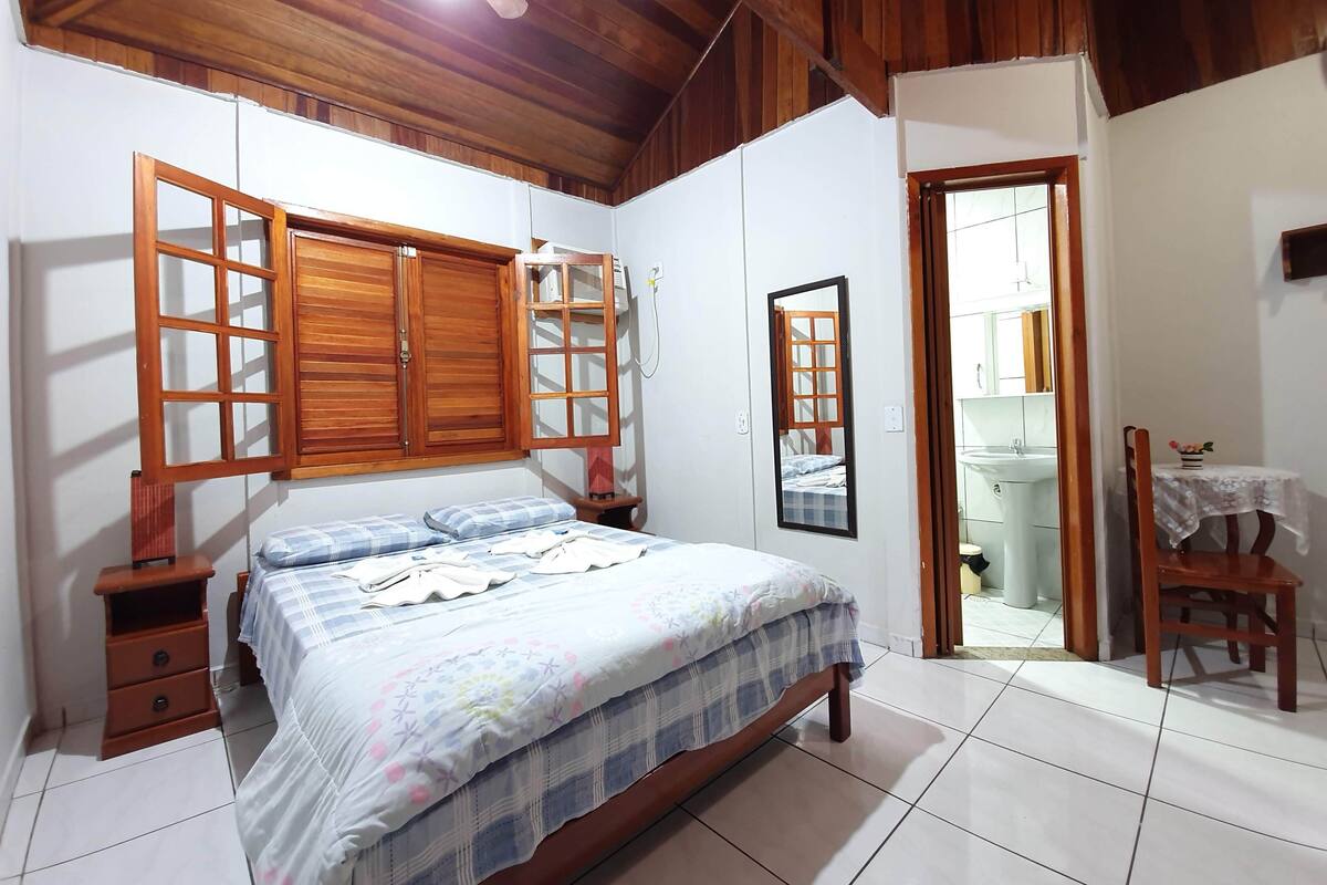 Quarto com cama, guarda-roupa de madeira, mesa pequena com cadeira e banheiro visível por uma porta aberta.