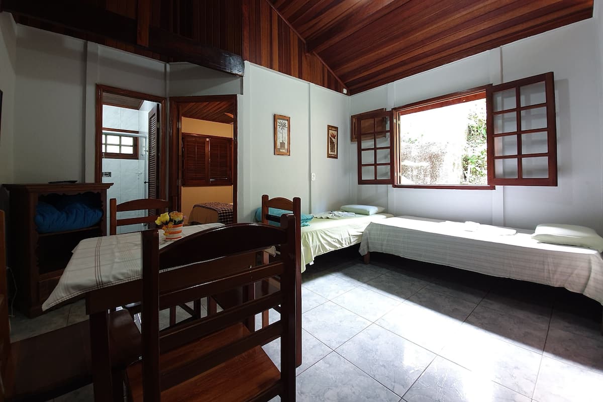 Quarto com cama, escrivaninha, cadeira, janela com persianas de madeira e ventoinha no teto.