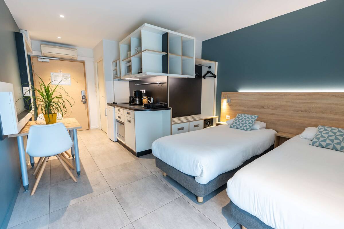 A modern hotel room with a kitchenette, two beds, a desk, and a potted plant.