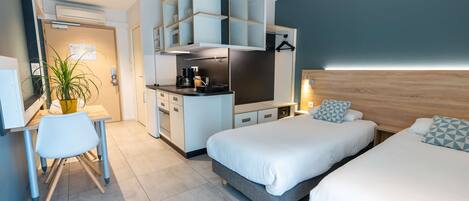 A modern hotel room with a kitchenette, two beds, a desk, and a potted plant.