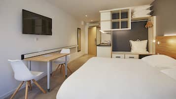 Une chambre d’hôtel moderne comprenant un lit, un bureau avec une chaise, un téléviseur fixé au mur et un dressing avec des étagères.