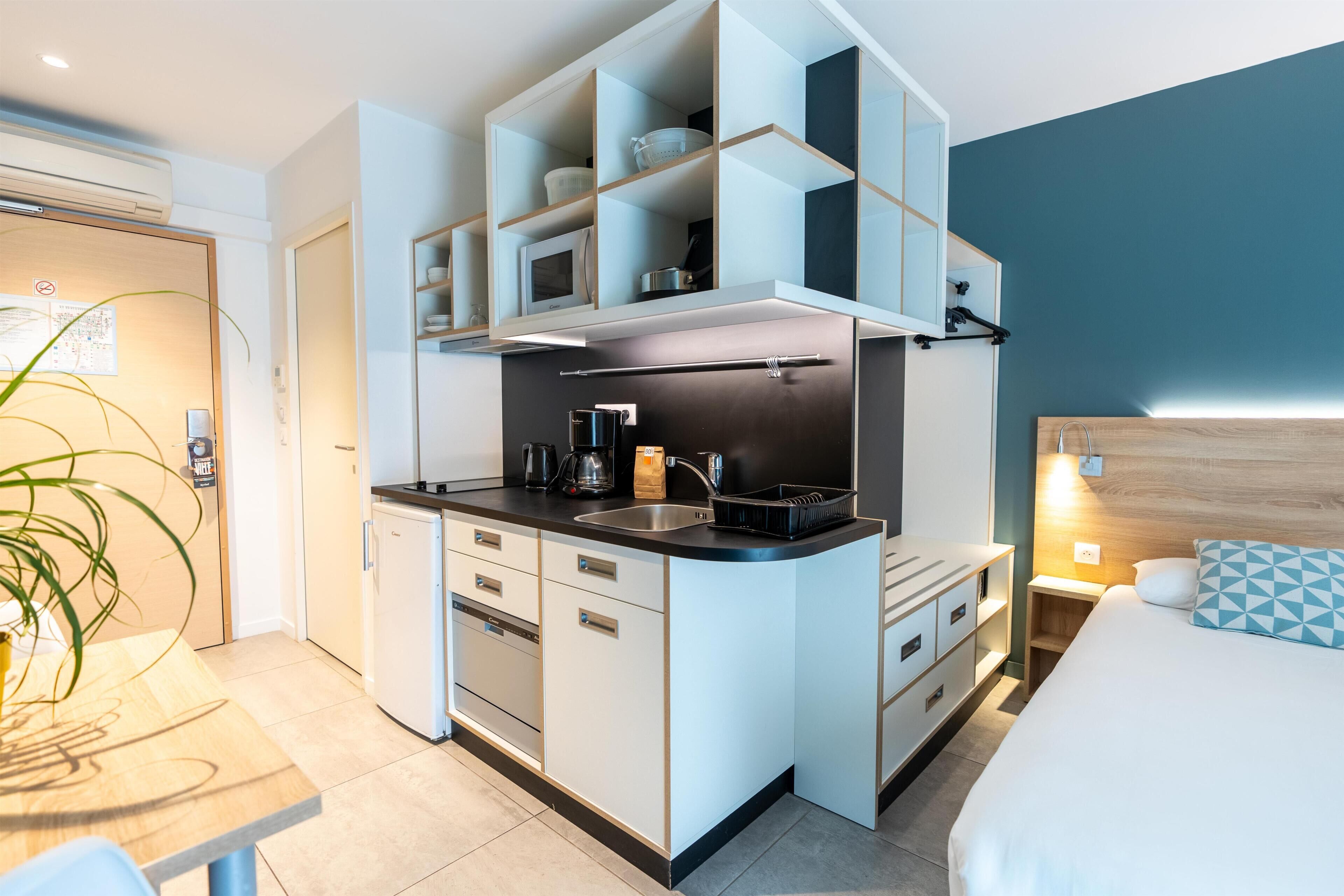 Una habitación de hotel compacta con kitchenette, una cama con almohada de diseño y una mesita de noche con lámpara.