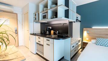Una habitación de hotel compacta con kitchenette, una cama con almohada de diseño y una mesita de noche con lámpara.
