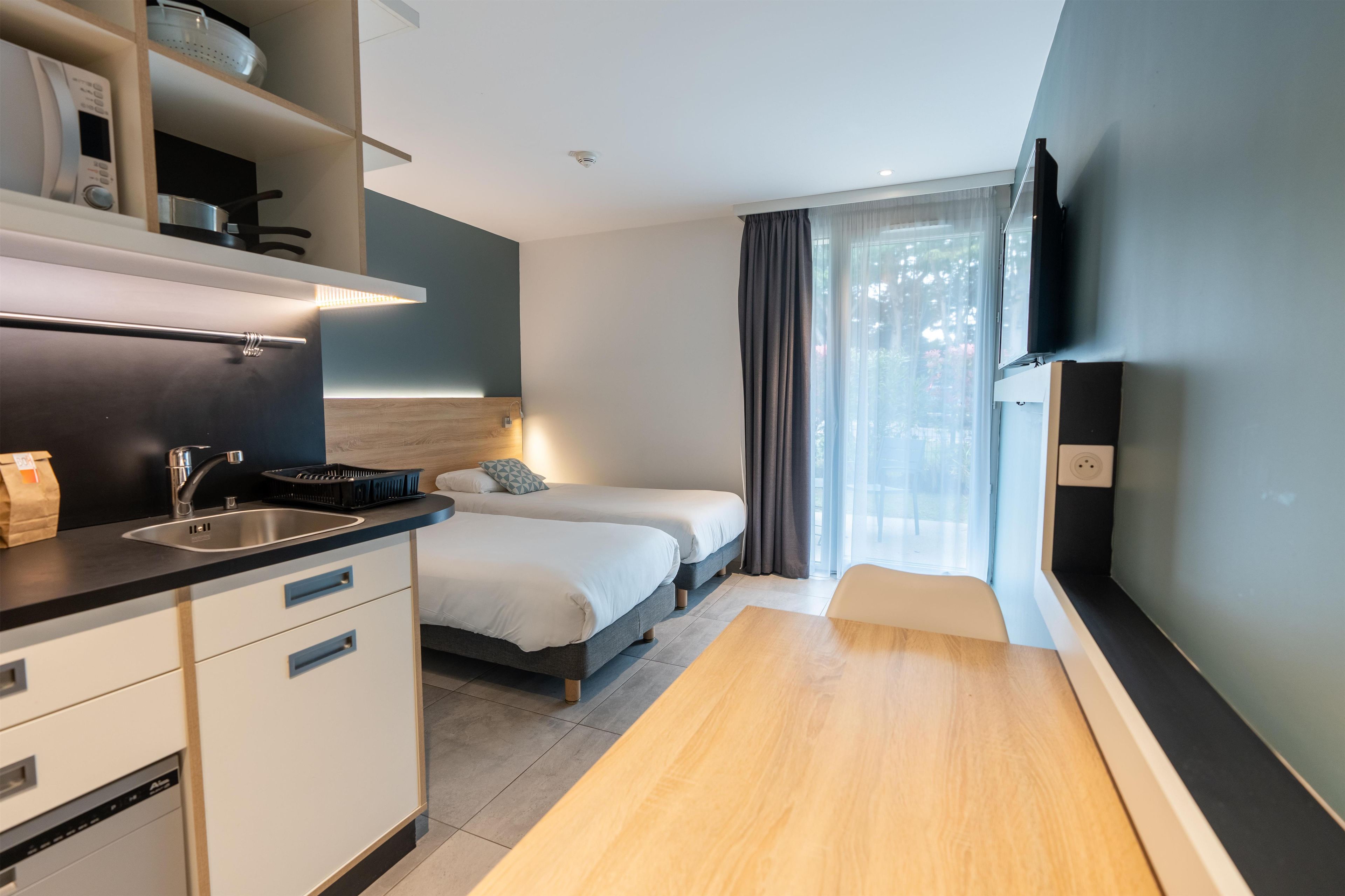 A modern hotel room with a kitchenette, a bed, a TV, and a window with curtains.