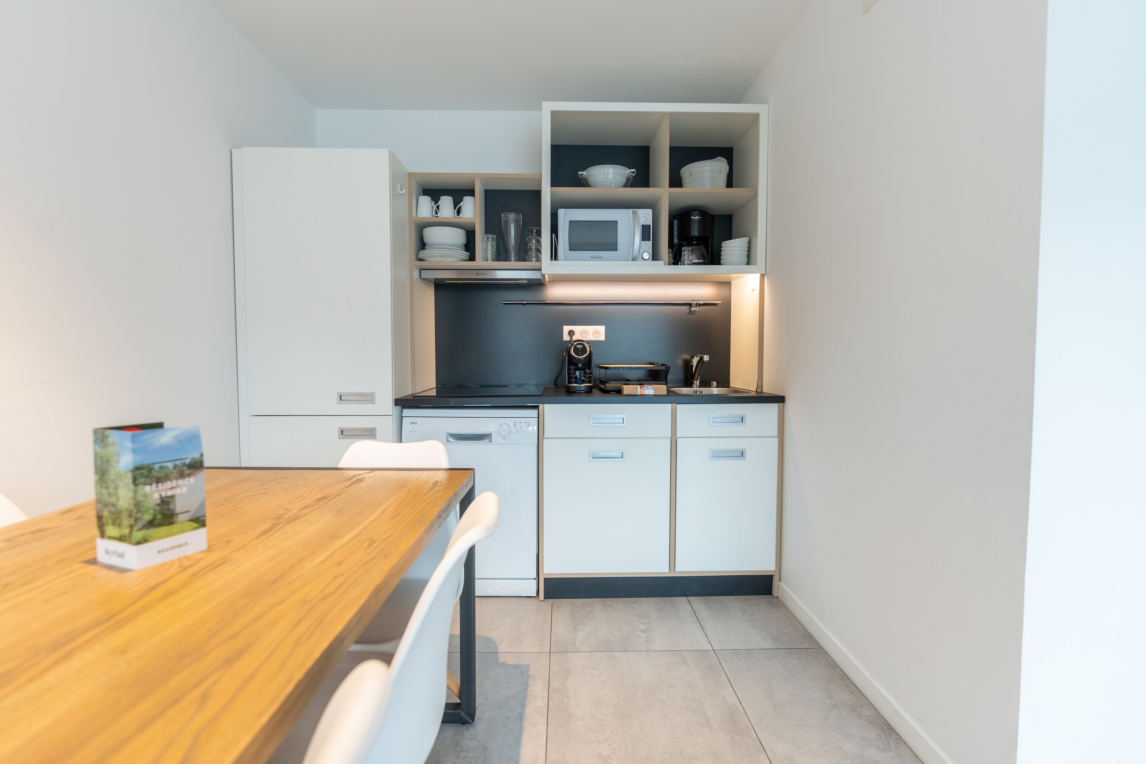 Cocina compacta con muebles blancos, microondas y cafetera. Hay una mesa de comedor con un libro encima.