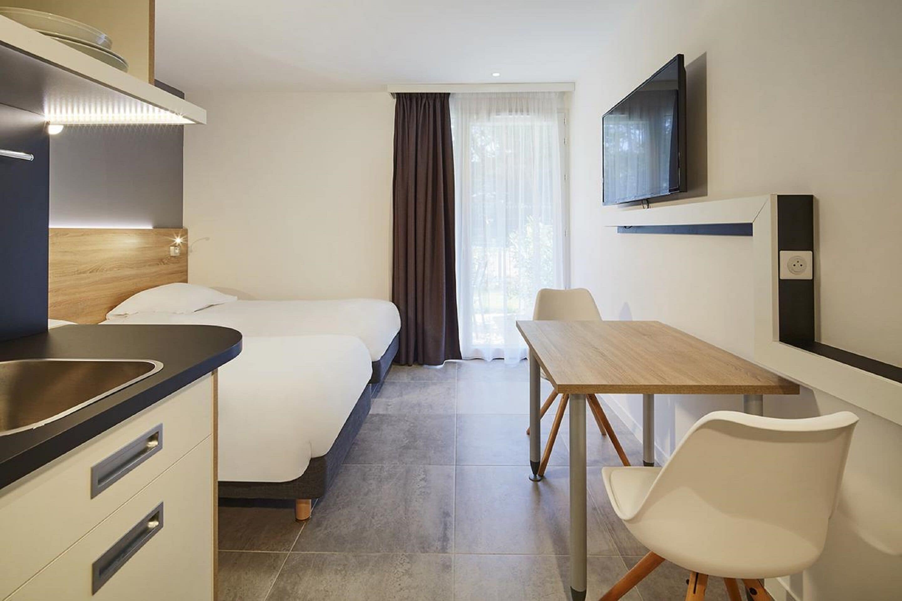 A hotel room with a bed, a desk, a chair, a TV, and a kitchen area.