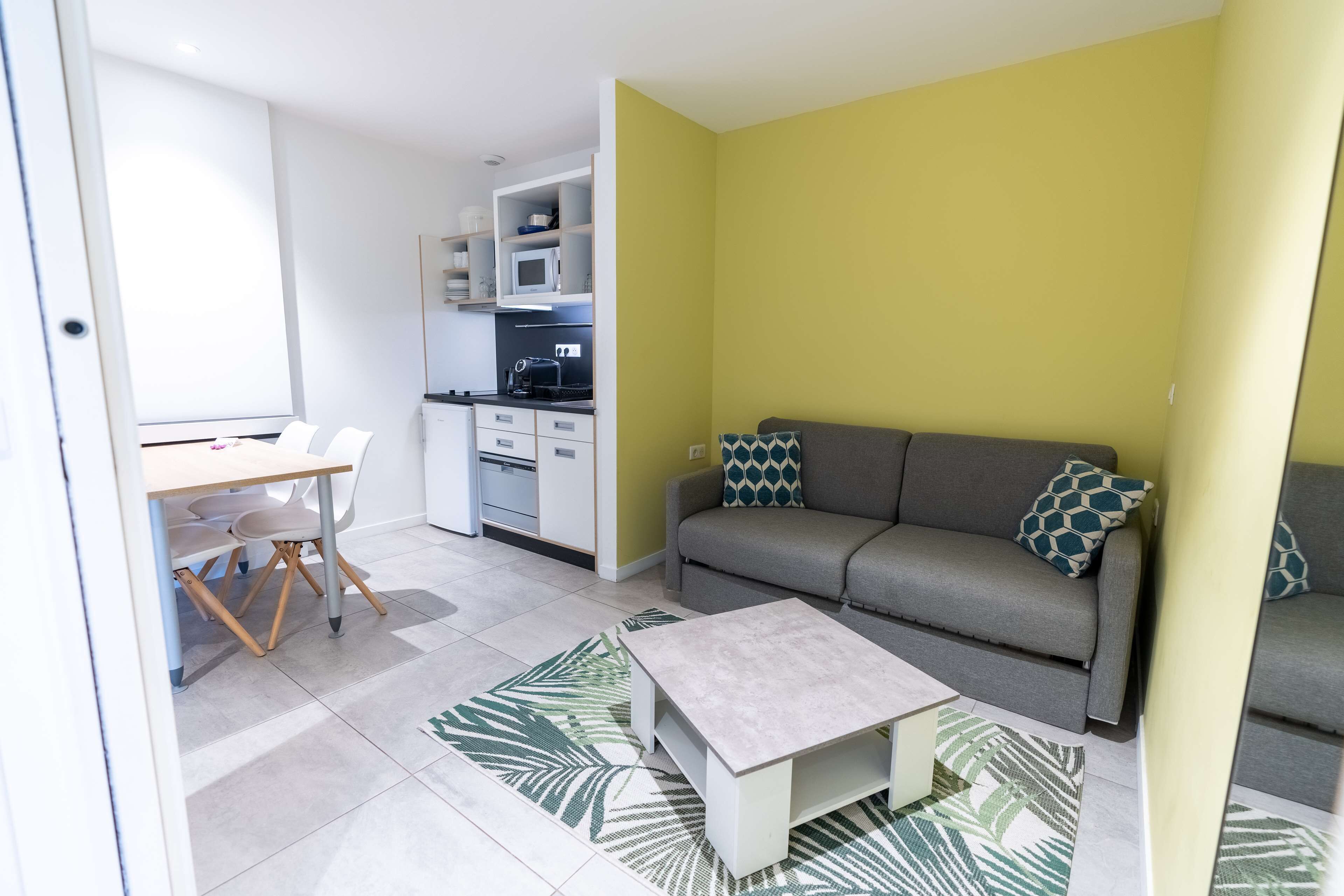 Un espace de vie compact comprenant un coin cuisine, un canapé gris agrémenté de coussins à motifs, une petite table basse blanche et un coin repas avec une table et des chaises blanches.