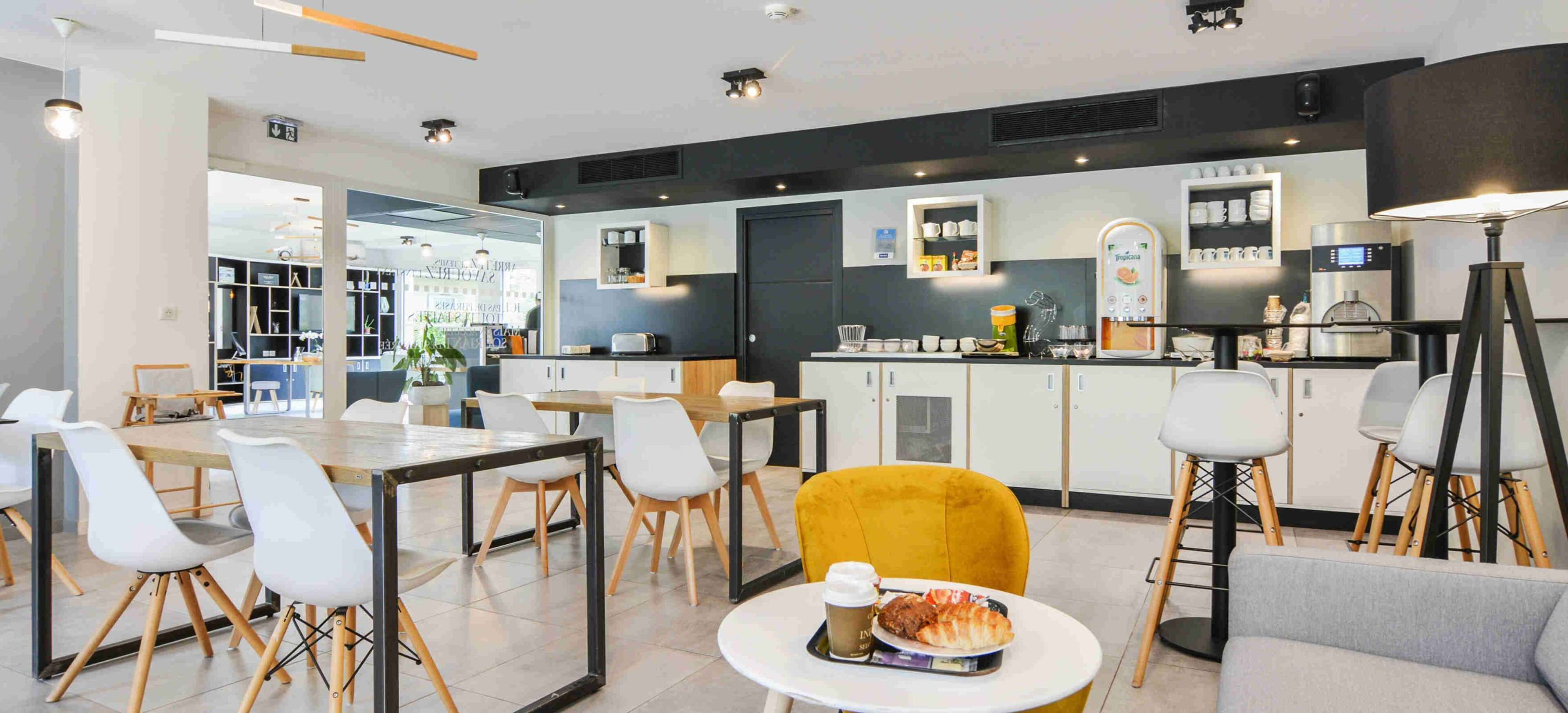 Un espace repas moderne avec une table centrale, des chaises jaunes et un canapé. Il y a également un coin cuisine équipé d’une machine à café et d’un réfrigérateur.