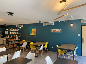 A modern dining area with wooden tables and colorful chairs, a kitchen area with shelves stocked with food items, and artwork on the walls. - Appart'hôtel Kyriad Résidence Cabriès - Plan de Campagne (Cabries)