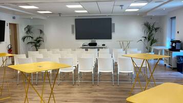 Un salón de conferencias con sillas blancas y mesas amarillas, una pantalla grande y plantas en macetas.