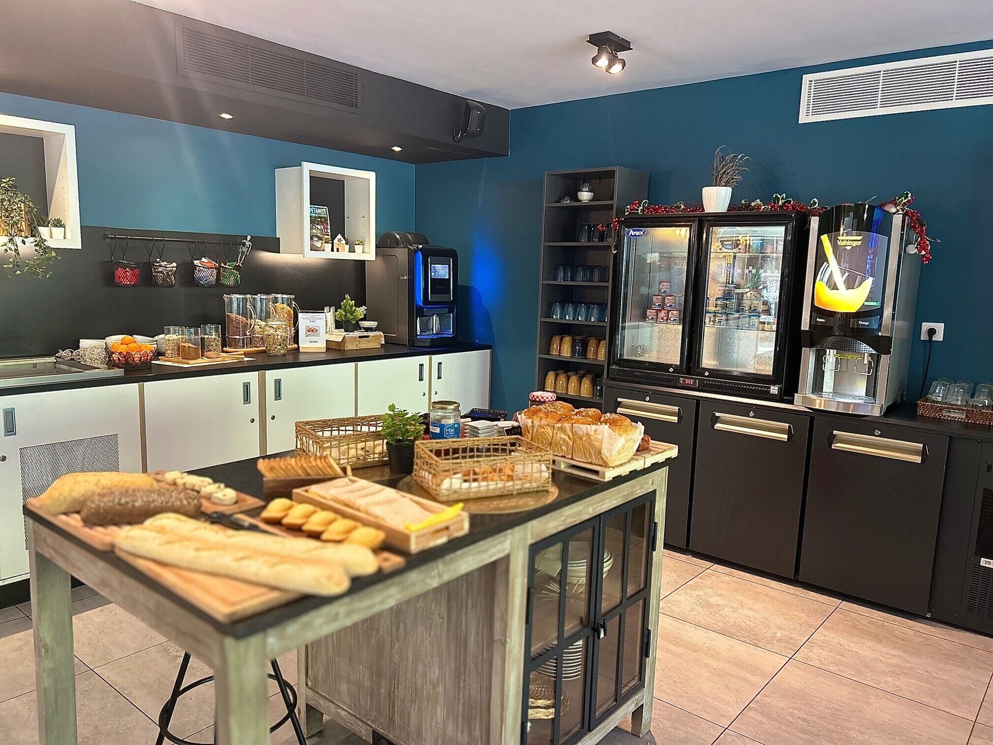 Eine Küchenzeile mit einem Tresen, auf dem Brot, Gebäck und eine Kaffeemaschine ausgestellt sind. Es gibt Regale mit Gläsern und einen Kühlschrank mit Kaffeemaschine.