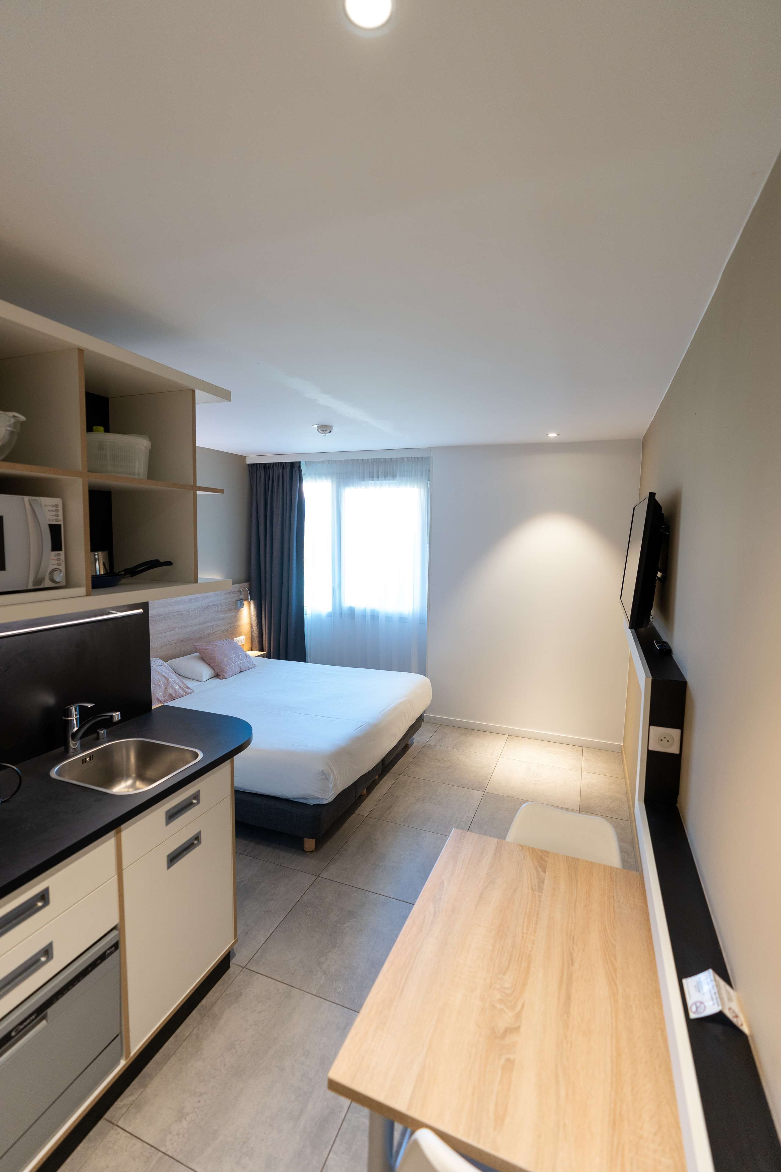 Une chambre d’hôtel de taille réduite, comprenant un lit, une petite kitchenette, une télévision et une fenêtre avec des rideaux.