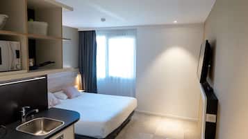 Une chambre d’hôtel de taille réduite, comprenant un lit, une petite kitchenette, une télévision et une fenêtre avec des rideaux.