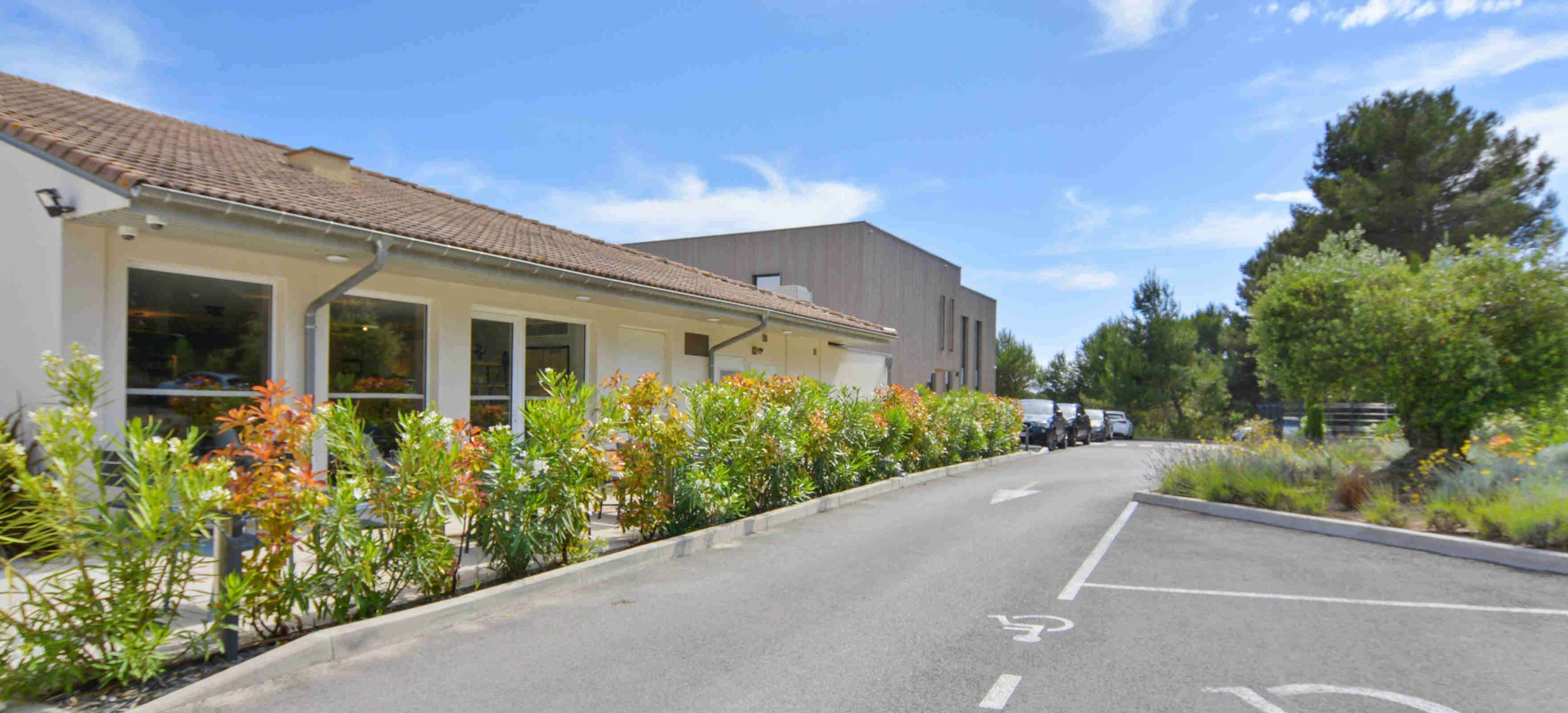 Un bâtiment à l’architecture moderne, un parking avec des places délimitées et une rangée de plantes fleuries le long du bord de la route.