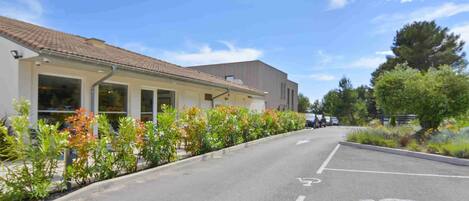 Un bâtiment à l’architecture moderne, un parking avec des places délimitées et une rangée de plantes fleuries le long du bord de la route.