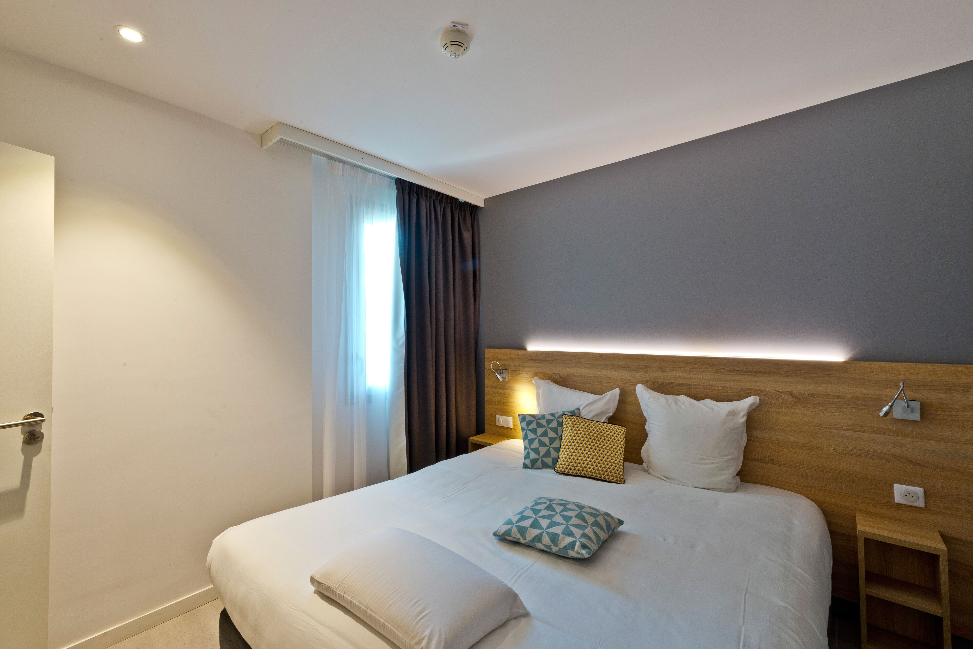 A modern hotel room with a large bed, a wooden headboard, and a bedside table with a lamp.