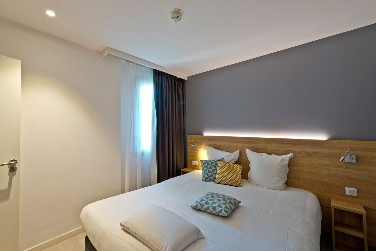 A modern hotel room with a large bed, a wooden headboard, and a bedside table with a lamp.