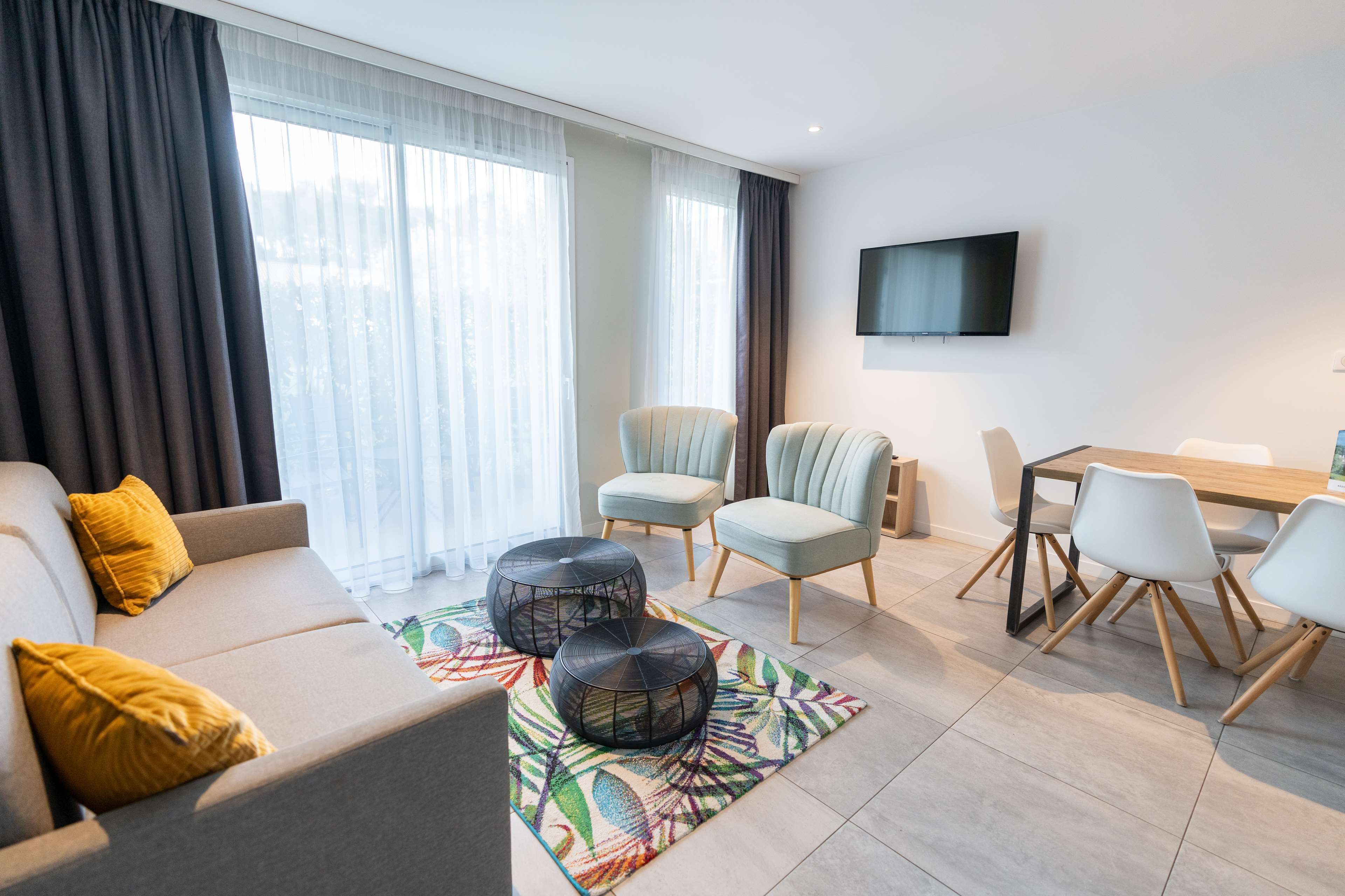 Un salon moderne comprenant un canapé, deux fauteuils, une table à manger avec des chaises, un téléviseur fixé au mur et une grande fenêtre avec des rideaux.