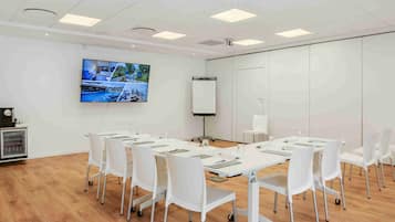 Une salle de conférence avec des tables et des chaises blanches, un écran de projection et un grand téléviseur fixé au mur.