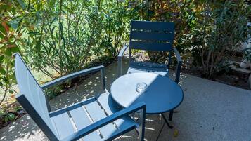 Zona de estar exterior de metal azul con una mesa y dos sillas, rodeada de vegetación y un edificio al fondo.
