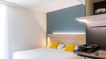 A modern hotel room with a bed, bedside table, and a kitchenette area.