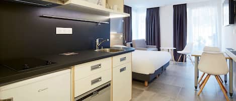 A modern hotel room with a kitchen area featuring a microwave, a dishwasher, and a sink. There is a bed with a gray headboard, a small desk with a chair, and a window with curtains.