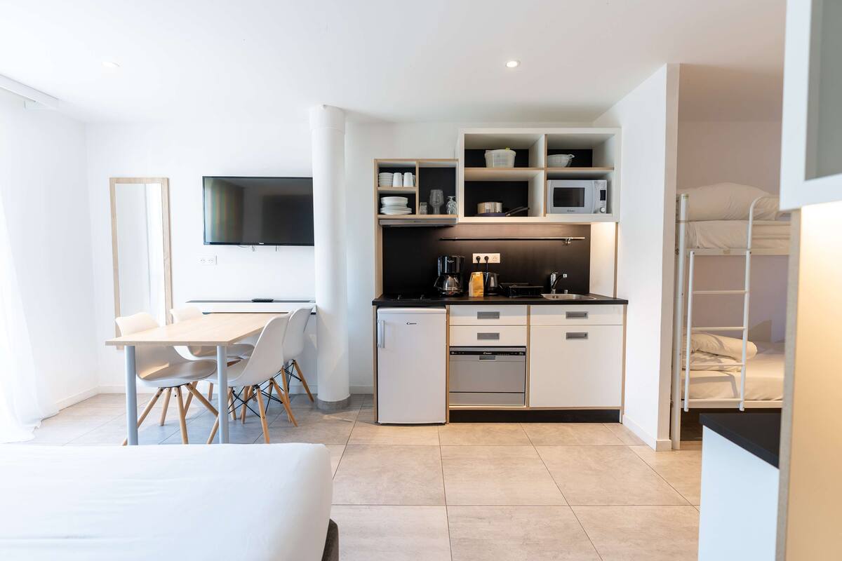 A compact, modern kitchenette with a built-in microwave, oven, and coffee maker. There is a small dining area with a table and chairs, and a TV mounted on the wall.